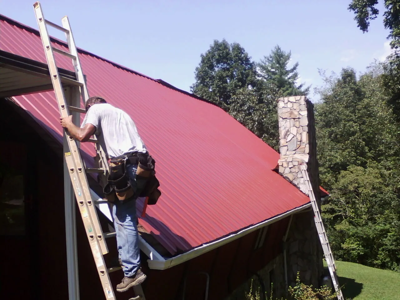 Roofer on a ladder beside a red metal roof and stone chimney — Soffit & Fascia Repair by Roofing Brothers in Bon Aqua